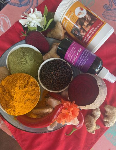 A colorful display of spices and ingredients for nutritional healing, including turmeric powder, black pepper, green powder, sliced beetroot, ginger, chili, garlic, flowers, and bottles of black seed and curcumin powder, on a red cloth. 999 Vedic