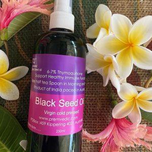 A bottle of Black Seed Oil with a spray cap lies on woven matting, surrounded by yellow and white frangipani flowers and green leaves. The label displays product details and website information.