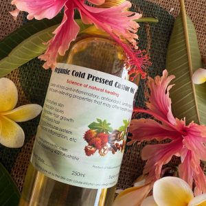 A bottle of organic cold pressed castor oil with a label detailing its benefits, surrounded by pink hibiscus flowers and green leaves, displayed on a brown textured surface.