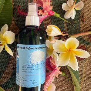 A bottle of cold-pressed organic magnesium spray sits on a woven mat, surrounded by tropical flowers and green leaves, with a description label explaining its uses for pain relief, stress, and fatigue.