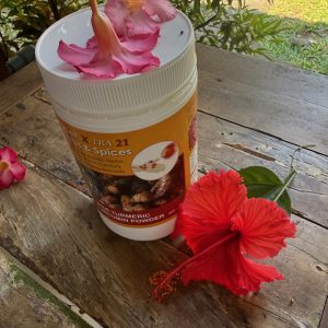 A white container of turmeric mix sits on a wooden table outdoors, topped with a pink flower. A large red hibiscus flower and green leaves rest beside it. Sunlight and greenery are visible in the background.