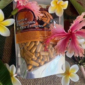 A resealable bag labeled Super Curcumin X-TRA 21 herbs filled with yellow capsules is surrounded by tropical flowers, including pink hibiscus and white plumeria, on a textured fabric surface.
