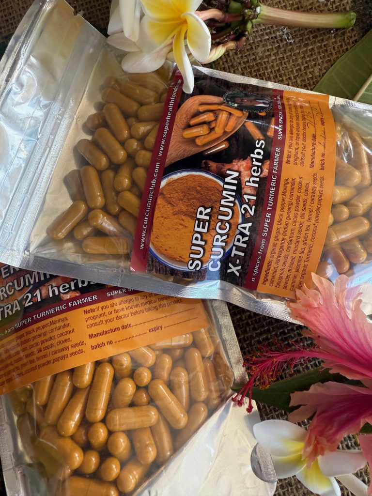 Two clear pouches labeled Super Curcumin X-TRA 21 herbs filled with yellow capsules, surrounded by tropical flowers and leaves on a natural woven surface.