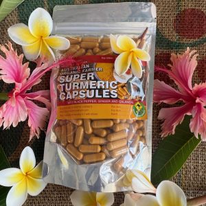 A foil pouch of Super Turmeric Capsules with black pepper, ginger, and galangal lies on a woven mat, surrounded by pink and white frangipani flowers and green leaves.